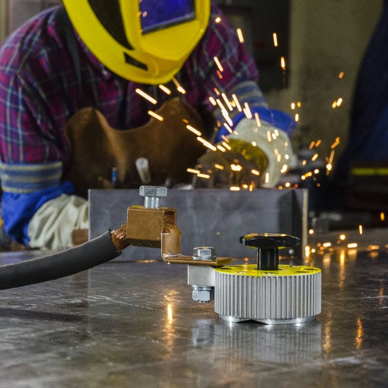Magswitch magnetic welding ground clamp attached to a metal surface with a welder working in the background