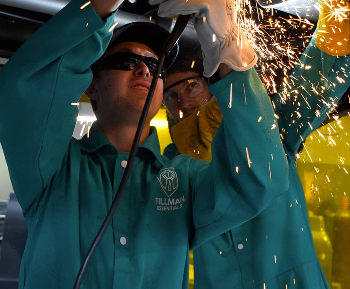 Worker wearing Tillman Essentials green welding jacket and safety glasses using a grinder with sparks flying
