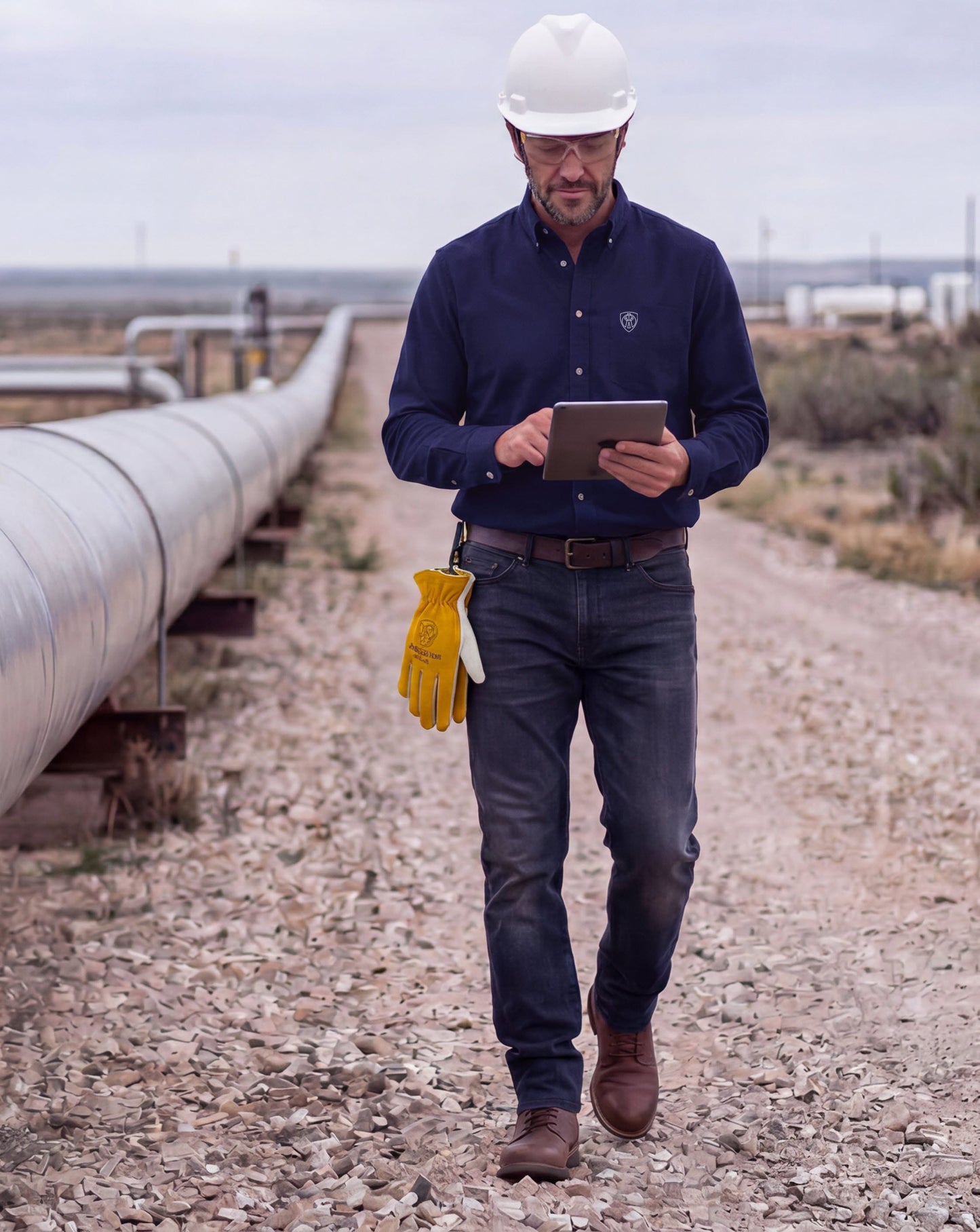 Worker in Tillman shirt and hard hat walking by pipeline using a tablet