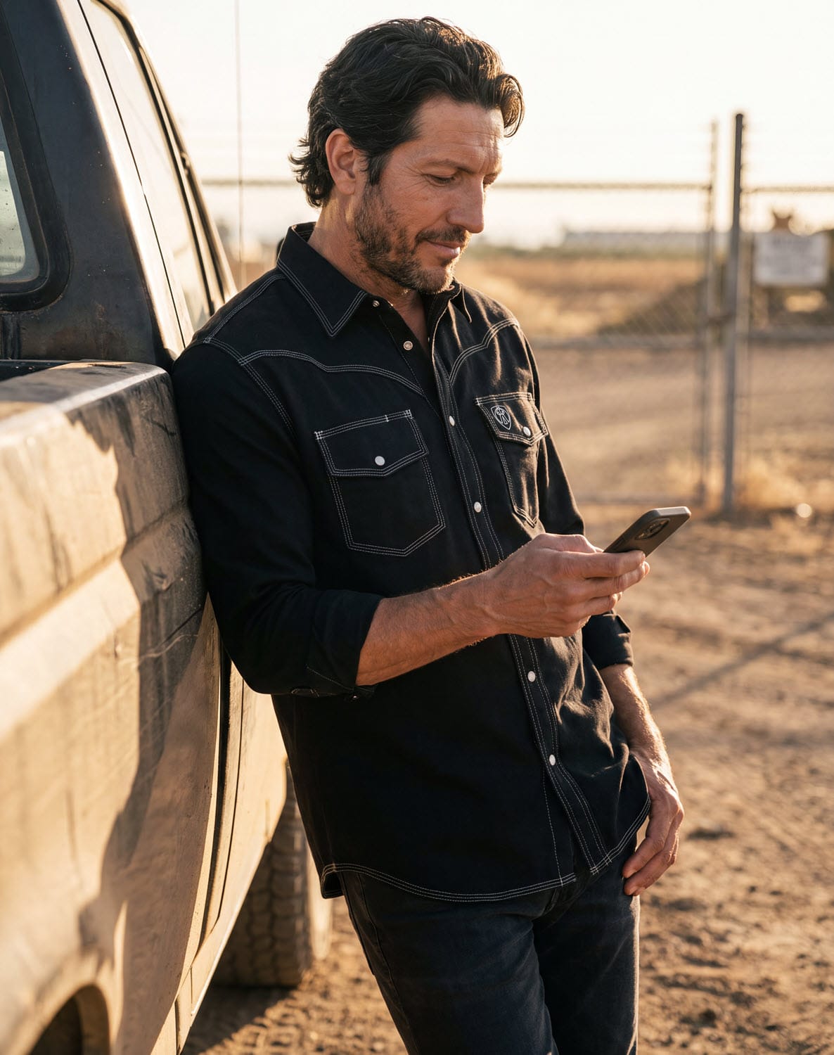 Man wearing a black Tillman western shirt with white contrast stitching leaning against a truck while using a phone
