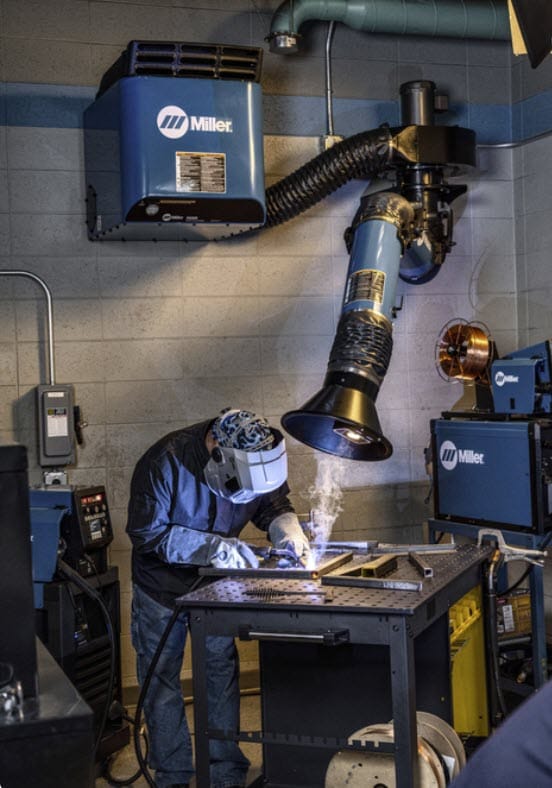 Welder wearing protective gear working at a workbench with a Miller fume extraction system
