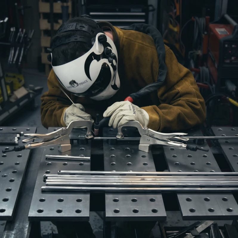 BuildPro welder wearing protective gear using a TIG torch on a metal table with locking clamps