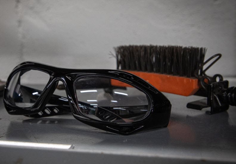 Black safety glasses with clear lenses next to a wire brush