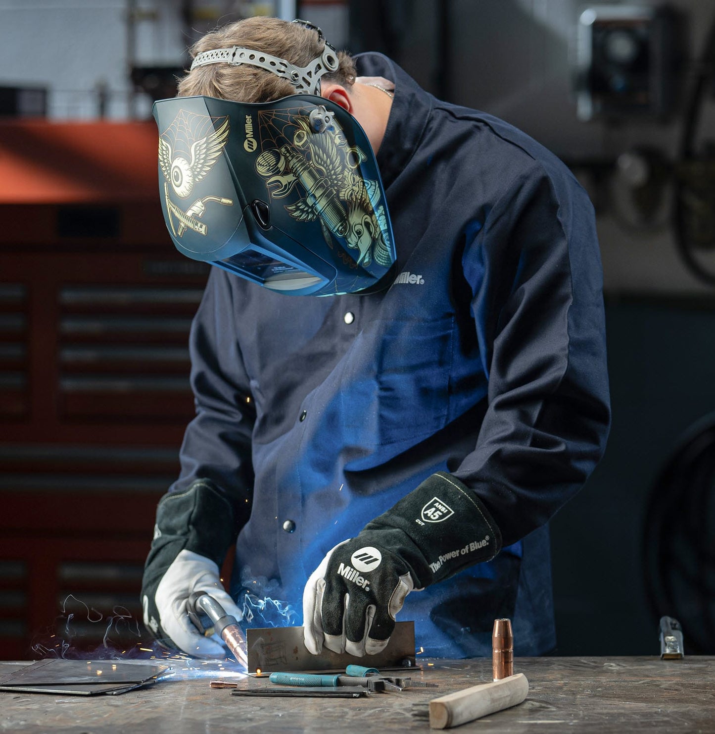 Welder wearing Miller helmet and gloves performing metal welding on a workbench